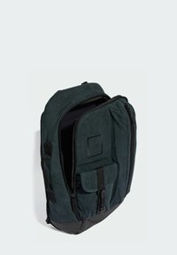 Zaino di tessuto verde scuro con due zip, tasca frontale e finitura texturizzata. Accenti neri alla base e patch del logo sul davanti.