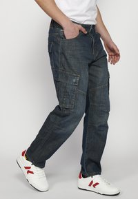 Kék farmer cargo nadrág több zsebbel, laza szabással, fakó textúrával és varrási részletekkel. Fehér sportcipőkkel párosítva, piros kiegészítőkkel.