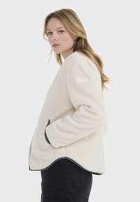 Chaqueta de forro polar beige con un corte relajado, bolsillos laterales y un ribete oscuro en contraste. Presenta una sutil etiqueta de la marca en el hombro izquierdo.