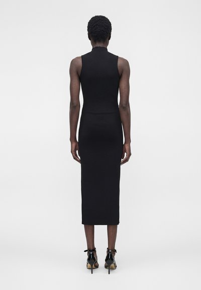 Άτομο με κοντά σγουρά μαλλιά που φοράει μαύρο midi φόρεμα χωρίς μανίκια και μαύρα σανδάλια με ψηλό τακούνι, στέκεται με την πλάτη προς τα εμπρός σε απλό φόντο.