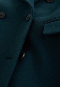 Gros plan sur un manteau en laine vert sarcelle foncé avec des boutons noirs et un détail de poche à rabat.