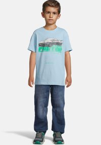 Šviesiai mėlynas medvilninis marškinėlis su grafiniu automobilio atvaizdu ir tekstu, derinamas su tamsiai mėlynomis džinsais ir pilkai žaliais sportiniais bateliais.