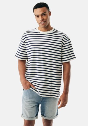 Junger Mann lächelt, trägt ein weißes und marineblau gestreiftes T-Shirt und hellblaue, hochgekrempelte Jeansshorts, steht vor einem einfarbigen Hintergrund.