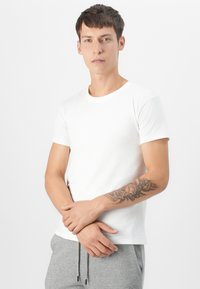 White cotton t-shirt with short sleeves, round neck, fitted design, worn with gray sweatpants. Notable floral tattoo on forearm.