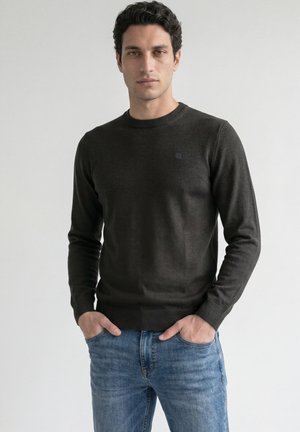 Mand med mørkt krøllet hår iført en sort langærmet sweater og blå jeans, står med hænderne i lommerne mod en ensfarvet baggrund.