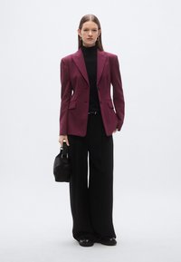 Bordeauxrood colbert over een zwarte coltrui, gecombineerd met zwarte pantalon met brede pijpen en een klein zwart handtasje. Gladde stof, getailleerde pasvorm.