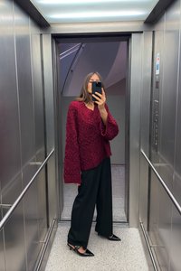 Burgundy adīta džemperis ar teksturētu rakstu, apkopots ar vaļīgām melnām biksēm un smailām melnām kurpēm. Spoguļojoša metāla lifta fons.