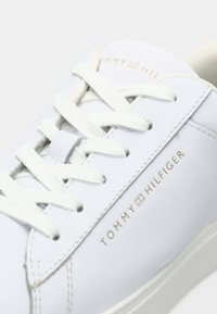 Gros plan d'une basket en cuir blanche avec des lacets blancs et le logo "Tommy Hilfiger" en lettres dorées sur le côté.