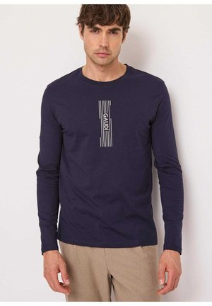 Uomo che indossa una camicia blu scuro a maniche lunghe con testo verticale "GAUDI" e design a righe, abbinata a pantaloni beige, in piedi davanti a uno sfondo semplice.
