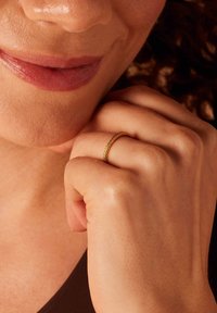 Goldring mit einem zarten, strukturierten Band, getragen an einer Hand, die gegen das Kinn ruht. Der Hautton erscheint warm und glatt.