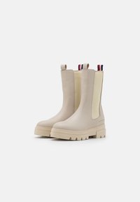 Botas de tobillo beige con paneles laterales elásticos, suelas de goma texturizada y lengüetas de tiro con acentos en rojo, blanco y azul. Acabado suave.