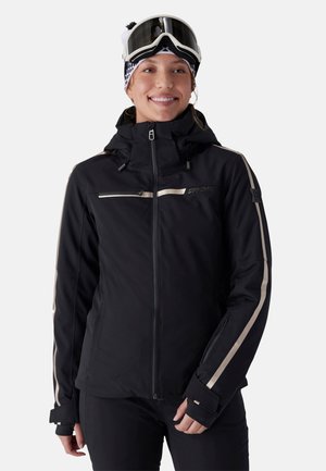 Femme portant une veste de ski noire avec des rayures beige, des lunettes de ski blanches sur le front et un bonnet en tricot, souriante et debout devant un fond blanc.