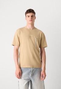 Beige, rövid ujjú póló, bal oldalon cipzáras zsebbel, puha anyagból készült, kerek nyakkivágással és diszkrét logóval.