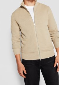 Mężczyzna ubrany w beżowy prążkowany sweter rozpinany na suwak, na biały shirt i czarne spodnie, stojący z rękami swobodnie opuszczonymi wzdłuż ciała.