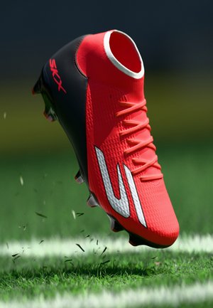 Chaussure de football rouge et noire avec des lacets, un logo blanc et des crampons soulevant de l'herbe sur un terrain vert près d'une ligne blanche.