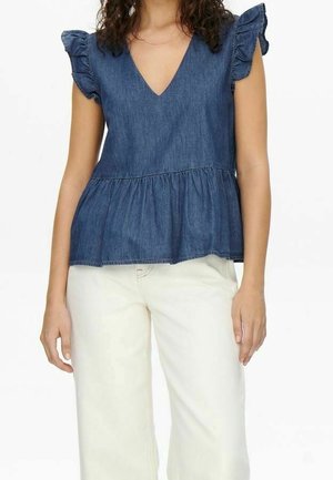 Kobieta ubrana w niebieski, bezrękawnik z denimu z falbanami na ramionach i z baskinką, zestawiony z białymi, wysokim stanem spodniami.