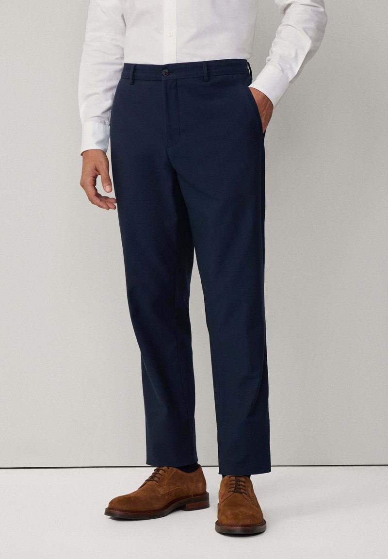 Pantalons de costume bleu marine à coupe droite, avec deux poches latérales et une fermeture à bouton, associés à des chaussures en daim marron et une chemise blanche.