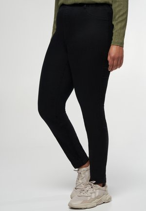 Personne portant un pantalon noir slim-fit, des baskets beige clair et un haut à manches longues vert olive, debout devant un fond uni.