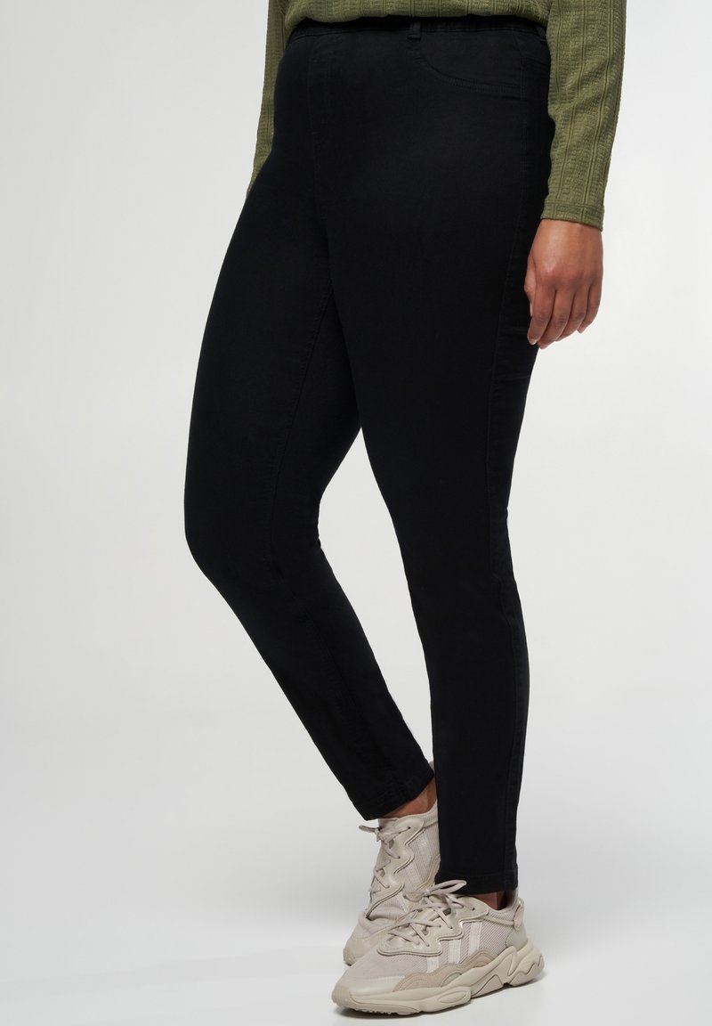 Personne portant un pantalon noir slim-fit, des baskets beige clair et un haut à manches longues vert olive, debout devant un fond uni.