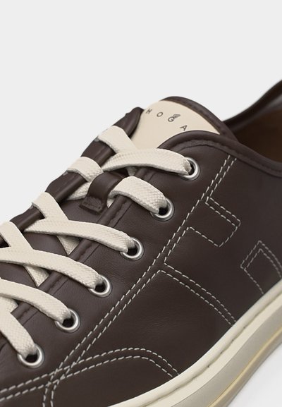 Primer plano de una zapatilla de cuero marrón oscuro con costuras blancas, cordones blancos, ojales de metal y una suela beige sobre un fondo neutro.