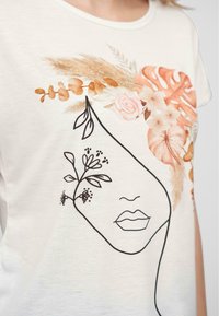 T-shirt blanc avec un design graphique représentant un visage rehaussé d'accents floraux dans des tons orange, rose et beige, entouré de noir.