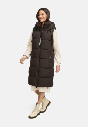 Veste longue et rembourrée de couleur marron foncé avec une capuche, fermeture éclair, poches latérales et cordons de serrage réglables. Portée par-dessus une robe de couleur claire d'une longueur jusqu'aux genoux.