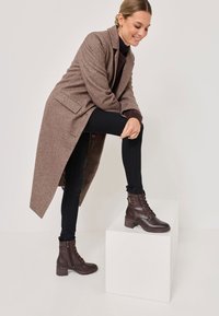 Cappotto di lana marrone con maniche lunghe, abbinato a jeans skinny neri e stivaletti alla caviglia marrone scuro con lacci e tacco spesso.