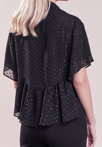 Top peplum noir à manches courtes, en tissu transparent avec un motif à pois subtil et un détail de taille froncée.