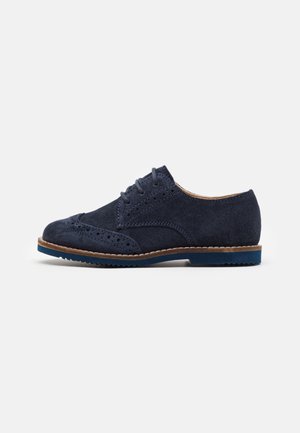Navyblå ruskinds oxford-sko med brogue-detaljer, brun kant, snørebånd og blå gummisål på hvid baggrund.