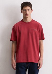 Rotes Baumwoll-T-Shirt mit kurzen Ärmeln, Rundhalsausschnitt und aufgedrucktem Text auf der oberen linken Seite; kombiniert mit dunklen Jeans. Weicher Stoff, lockere Passform.