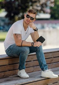 Mladý muž v bílem tričku sedí na dřevěné lavičce, má na sobě černé sluneční brýle, modré džíny a bílé tenisky, v ruce drží černou peněženku.