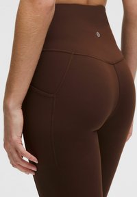 Pruunid kõrge vöökohaga leggingud, millel on sile tekstuur, küljetasku ja peened õmbluse detailid, mis lisavad struktuuri.