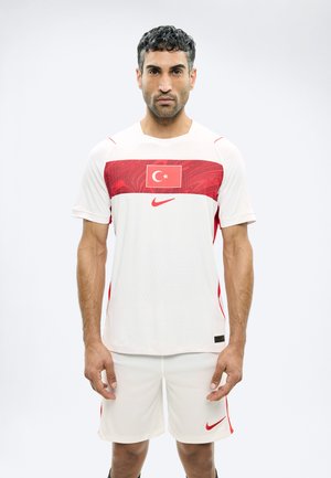 Άνδρας που φοράει αθλητική φανέλα και σορτς σε λευκό και κόκκινο με τη σημαία της Τουρκίας και το λογότυπο της Nike, στέκεται μπροστά σε απλό λευκό φόντο.