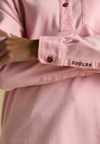 Hele roosa pika varrukaga särk, millel on nööpidega krae, pruuni nööpiga mansetil ja 'JOULES' tikand õmbluse lähedal.