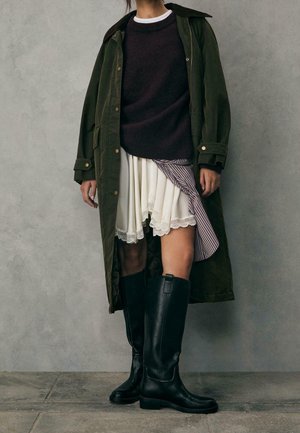 Person iført en mørkegrøn lang frakke, sort sweater, hvid lagdelt nederdel med blonderkant og sorte knæhøje støvler, stående mod en grå væg.