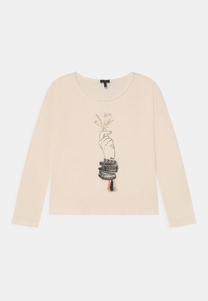 Chemise beige à manches longues avec un motif main tenant de petites fleurs et plusieurs bracelets tissés colorés avec des pompons au poignet.