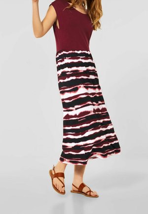 Femme portant une robe sans manches bordeaux avec une jupe tie-dye horizontale noire, blanche et rouge et des sandales plates marron, debout sur un sol gris.