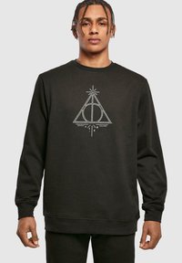 Svart hettegenser med hvit grafisk design av en trekant med en sirkel og linjer, tekst "Deathly Hallows" under, ribbestrikkede mansjetter og fald.