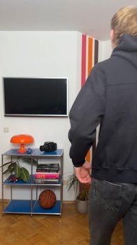 Person i sort hættetrøje vendt mod et TV monteret på en hvid væg, nær en blå metalhylde med en basketball, bøger, kamera, lampe og planter.