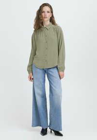 Sieviete ar viļņainiem matiem, valkājot olīvu zaļu pogājamu blūzi, gaišo zilo platā kāju džinsu bikses un melnus zābakus ar papēžiem uz vienkārša fona.