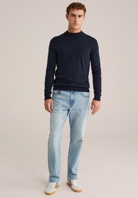 Man med lockigt hår iförd en marinblå långärmad tröja, ljusblå jeans och vita sneakers mot en beige bakgrund.