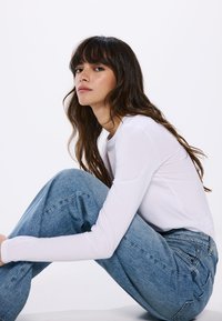 T-shirt bianco a maniche lunghe e jeans azzurri a vita alta. La t-shirt è aderente, mentre i jeans hanno una vestibilità rilassata e ampia con cuciture visibili.