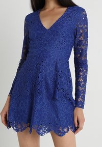 Robe en dentelle bleu roi avec manches longues, encolure en V et ourlet festonné. Caractérisée par des motifs floraux et une silhouette évasée.