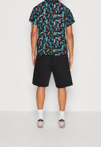 Korte mouwen, zwart shirt met kleurrijke geometrische patronen; gecombineerd met zwarte denimshorts. Casual schoenen met accentkleuren.