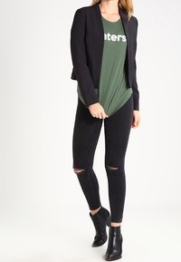 Femme portant un blazer noir, une chemise verte avec un texte blanc partiel, un jean skinny noir déchiré et des bottines noires, debout devant un fond blanc.