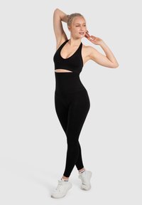 Set sportivo nero composto da un reggiseno a halter con apertura e leggings a vita alta. Modello con sneakers bianche, tessuto liscio.