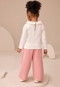 Haut blanc à manches longues avec col à volants, dos boutonné ; pantalon rose à jambes larges et chaussures blanches. Matériaux simples et doux avec une coupe décontractée.