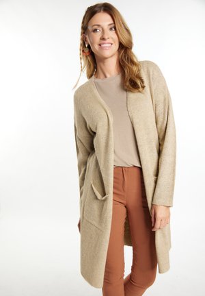 Beige Cardigan mit weicher Textur, offener Vorderseite, zwei Taschen und langen Ärmeln, kombiniert mit einem hellbraunen Oberteil und rostfarbenen Hosen.