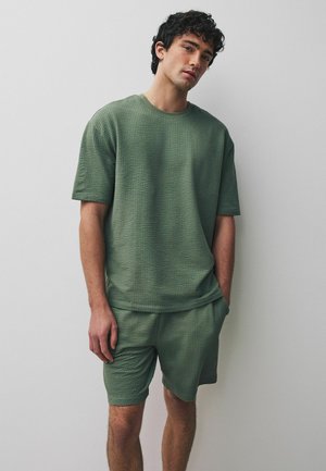 Young man standing against a plain wall wearing textured olive green short-sleeve shirt and matching shorts with hands in pockets.