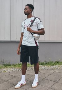 Lys blå sportsjersey med maroon detaljer, sorte shorts, hvide socks og røde stribede sneakers. Sølv crossover taske. Stående udendørs.